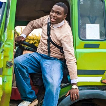 A smiling young man in casual clothing sits in a green truck in Kaduna, Nigeria.